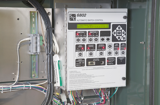Uma unidade de Controle de Chaves 6802 montada na parede. A unidade está no lado direito da foto, enquanto o lado esquerdo mostra fios presos à parede que leva à unidade. Os fios também se projetam da parte inferior da unidade e para fora da moldura da imagem. A unidade de Controle Controle Automático de Interruptores 6802 tem um logotipo da S&C no canto superior esquerdo do painel frontal. O painel frontal tem também vários botões de touchpad rotulados; O &quot;Botão 1&quot; e o &quot;Botão 2&quot; são legíveis, todos os outros textos são muito pequenos para ler. Em alguns botões, luzes vermelhas estão acesas. 