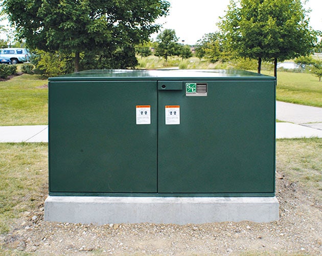 front view of an installed P M H pad-mounted gear for outdoor distribution, 14.4 kV and 25 kV with roof and doors closed on a concrete pad with grassy field and trees in the background PMH Pad-Mounted Gear