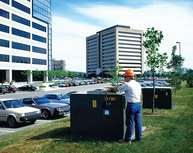 front view of an installed near tall office buildings near a parking lot to the left of the P M H pad mounted gear for outdoor distribution, 14.4 kV and 25 kV with a lineman using a penta latch to open the right door. The sky is blue closed on concrete pad with grassy field and trees in the background PMH Pad-Mounted Gear